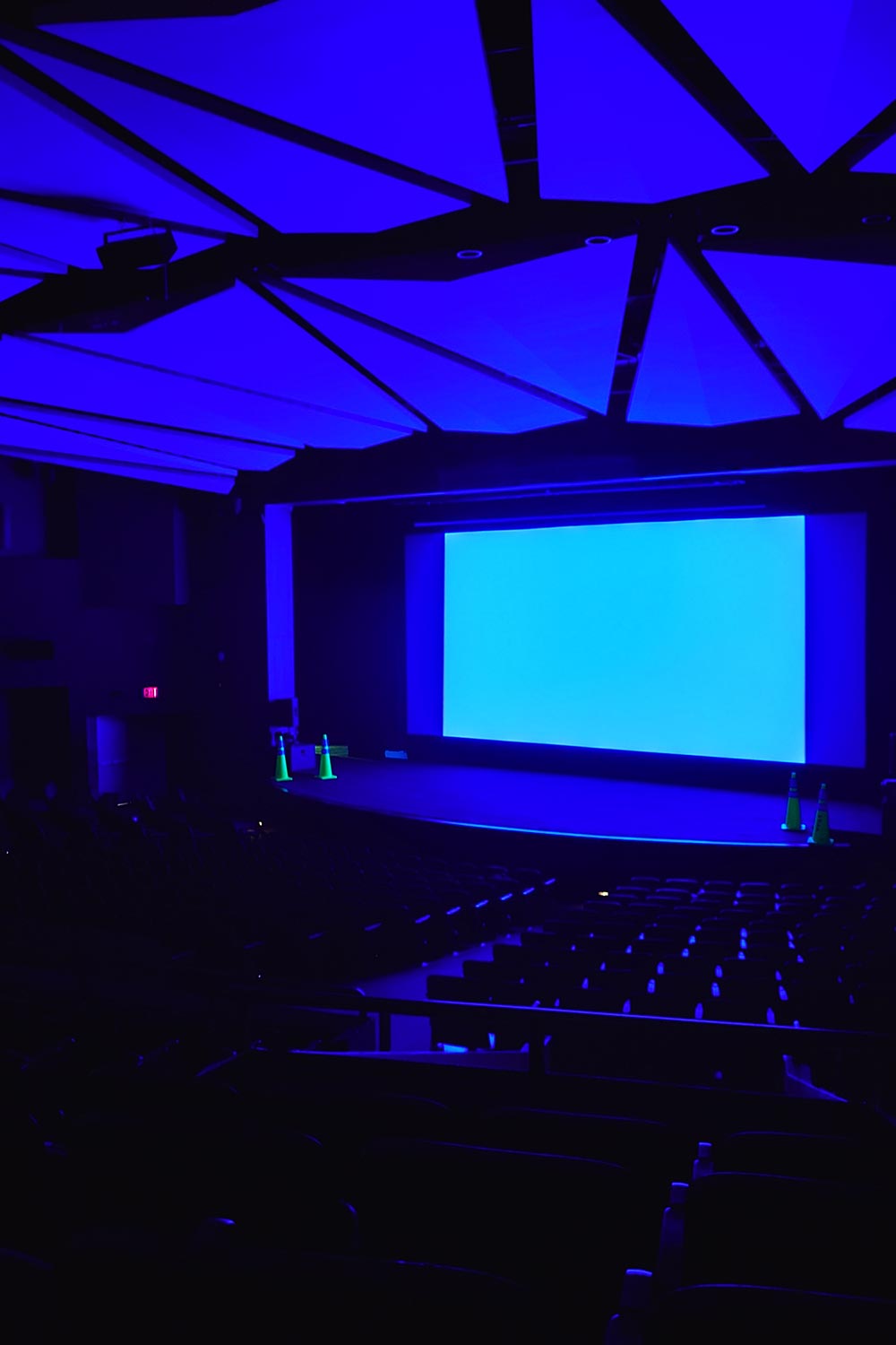 Dark theater with blue-lit ceiling and screen, green safety cones near stage