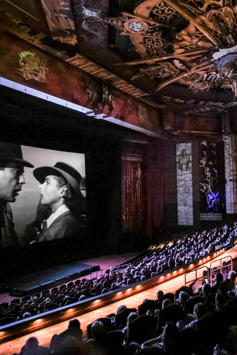 Audience watching a black and white classic film in an ornate theater with a decorated ceiling