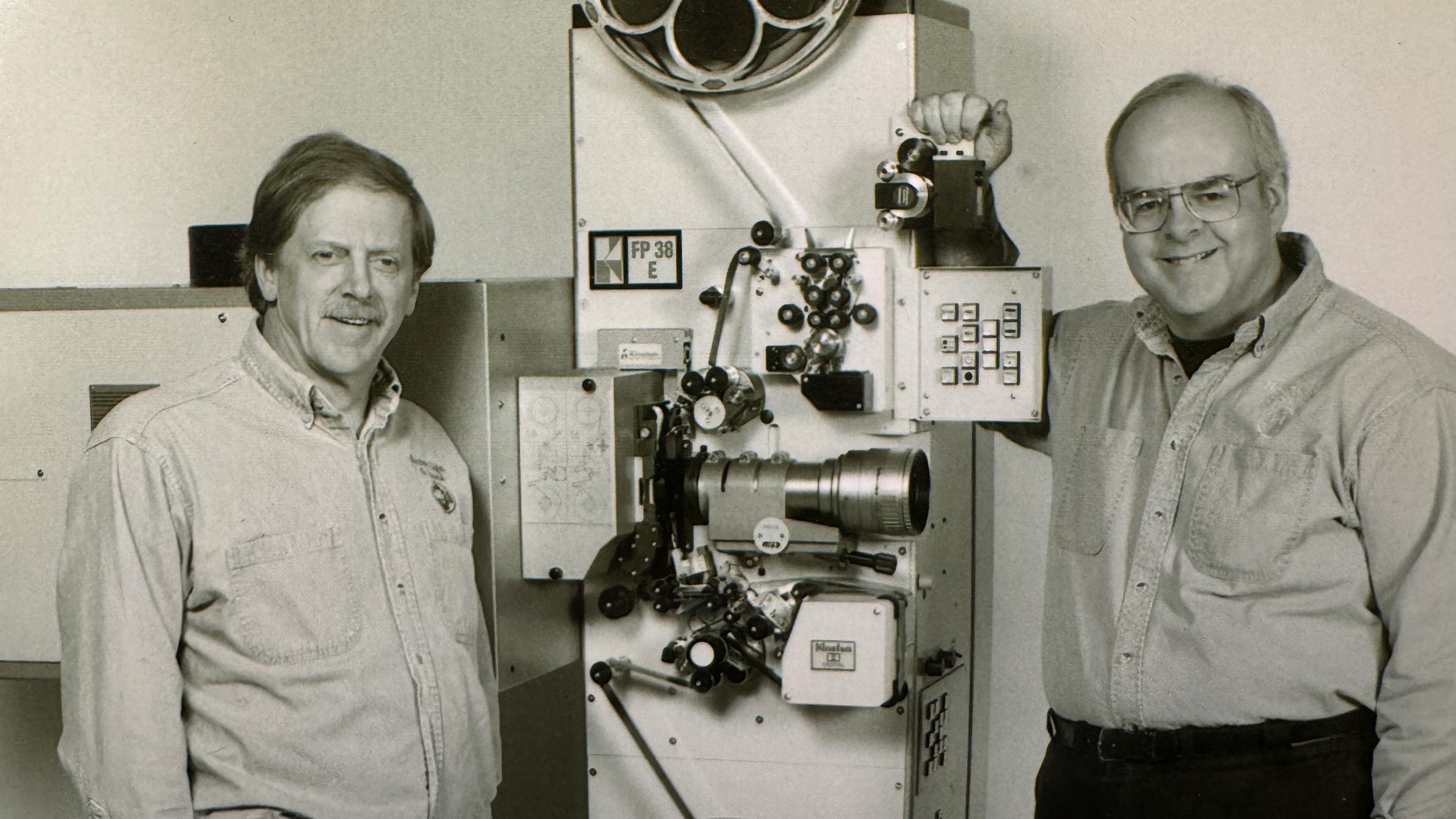 Two smiling men posing beside an FP 38 E film projector unit