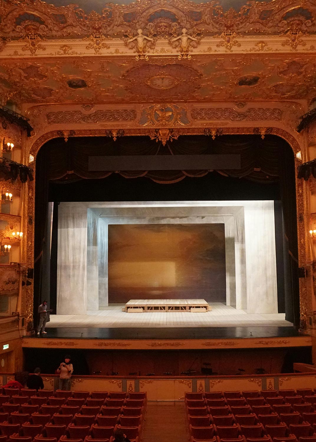 A grand theater stage with an ornate ceiling and a minimalist set design, viewed from the audience seating area.