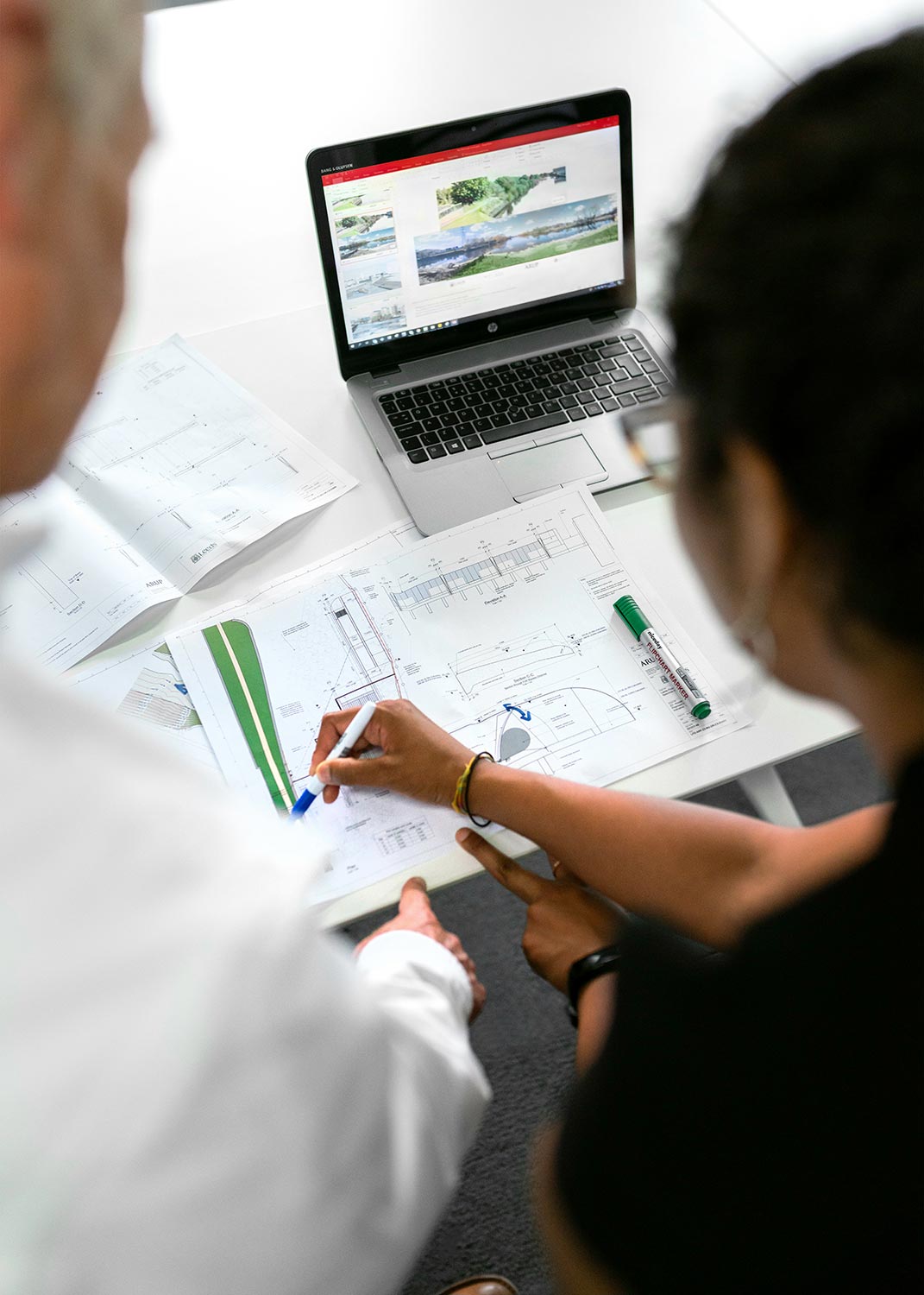 Two people reviewing architectural plans on a table with a laptop showing images