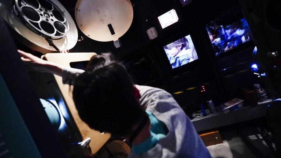 A person operating a vintage film projector in a dimly lit room with multiple screens displaying scenes from a film.