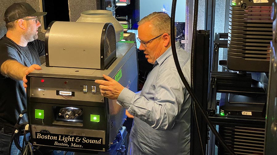 Two men carefully installing a large professional projector in a technical workspace, surrounded by high-tech equipment.