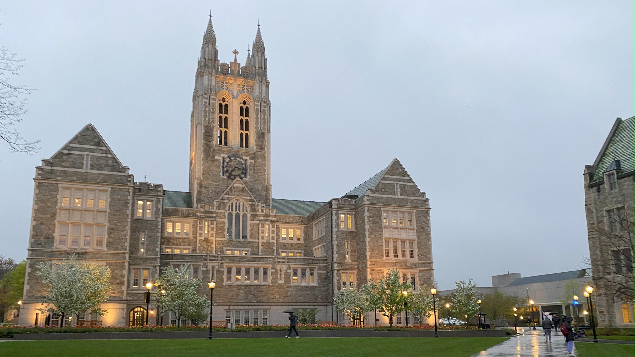 Gothic-style stone building at Boston College illuminated at dusk with people walking nearby