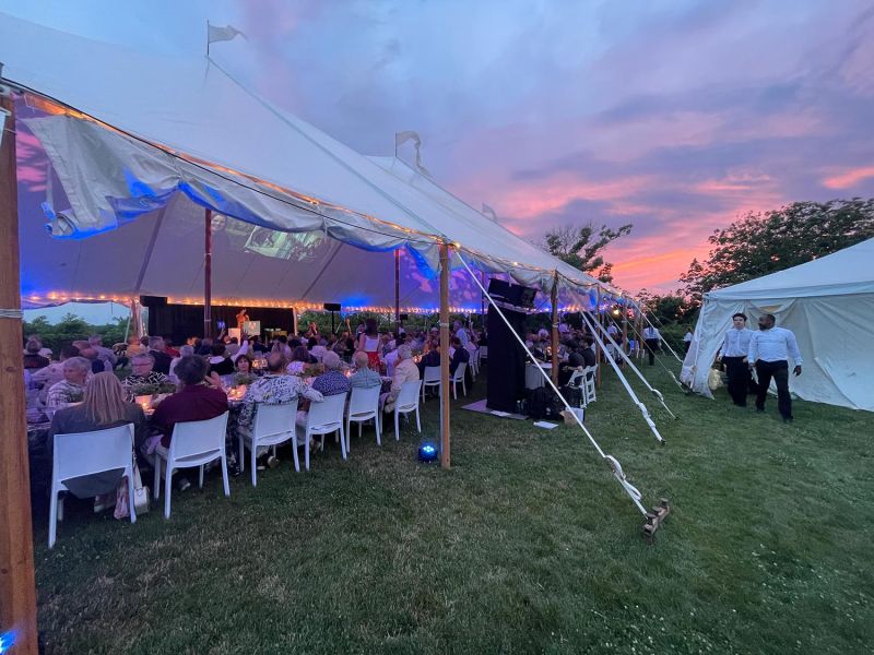 Outdoor tented dinner event at sunset with guests seated and speakers on stage