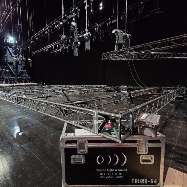 Stage setup in progress with metal trusses, suspended lighting fixtures, and a Boston Light & Sound equipment case on a glossy black stage floor